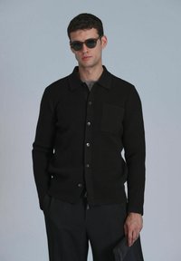 Schwarze gerippte Strickjacke mit Kragen, Gesäßtasche und Knopfverschluss, getragen über einem grauen Hemd und kombiniert mit schwarzen Hosen.