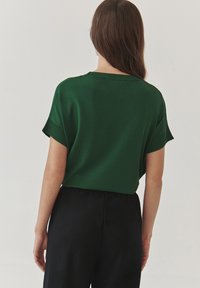 Grüner kurzärmeliger Pullover mit überschnittenen Schultern, lässigem Schnitt und glatter Textur. Getragen mit schwarzen Hosen. Einfaches, minimalistisches Design.