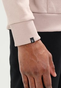 Primo piano di una mano di carnagione scura accanto al polsino di una manica a coste beige con un piccolo etichetta di stoffa nera attaccata.