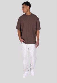 Brown oversized t-shirt, short sleeves, round neckline. Paired with white distressed pants and white sneakers. Model standing sideways.