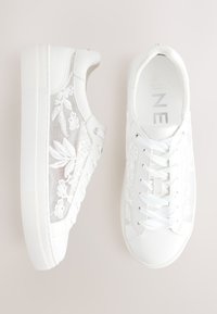 Zapatillas blancas con cordones, con paneles de malla bordados con flores y suelas gruesas de goma, mostradas desde vistas lateral y superior.