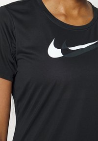 Camisa atlética negra de manga corta con logos en capas en blanco y negro en el pecho, usada por una persona de piel marrón.