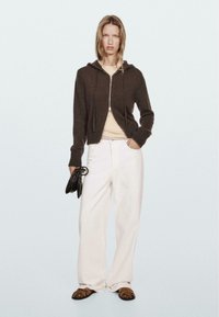 Brown zip-up hoodie, cream top, white wide-leg pants, and brown sandals. Model holds a black handbag, with straight hair and neutral background.