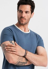 Niebieska koszulka z krótkim rękawem z kontrastowym białym dekoltem, o miękkiej teksturze i luźnym kroju. Wyraźne tatuaże i nałożone bransoletki na ramionach.