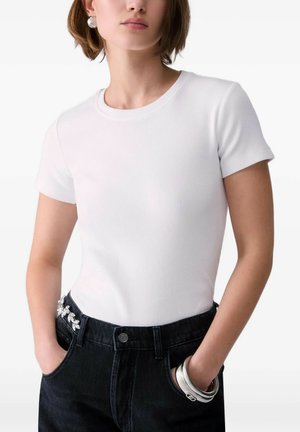Donna che indossa una semplice maglietta bianca a maniche corte infilata nei jeans neri con braccialetti e orecchini d'argento, mani in tasca.