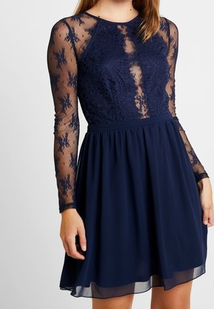 Kvinde iført en mørkeblå kjole med gennemsigtige blonder lange ærmer og blonder korsage samt et ensfarvet, flydende skørt.