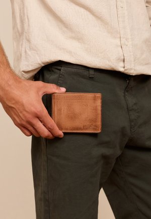 Braune Lederbrieftasche, die von einer Hand gehalten wird, rechteckig mit glatter Textur, mit gestickten Kanten und einem kleinen Logo auf der Vorderseite.