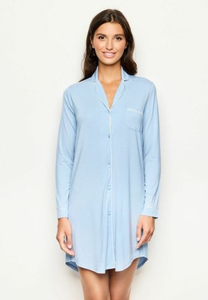 Donna che indossa una camicia da notte blu chiaro con maniche lunghe e bottoni, rifinita con piping bianco e una tasca sul petto, in piedi su uno sfondo neutro.