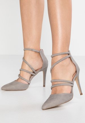 Zapatos de tacón alto de ante taupe delgados con puntera puntiaguda y múltiples finas correas en el tobillo sobre los pies desnudos, usados por una persona de pie.