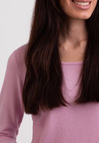 Helles rosa Sweatshirt mit weicher Textur und lässigem Schnitt, ausgestattet mit einem quadratischen Ausschnitt. Die Haare sind lang und dunkel und bedecken teilweise die Schulter.