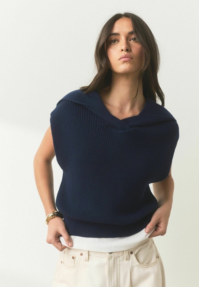 Femme aux longs cheveux foncés portant un pull en maille sans manches bleu marine par-dessus un haut blanc et un pantalon clair, posant avec les mains près des poches.