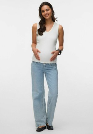 Trudna žena u bijelom topu bez rukava i svijetloplavim trapericama, smiješi se i nježno drži trbuh na jednobojnoj pozadini.