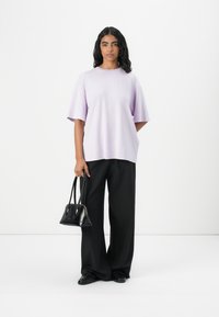 Tricou oversized lila cu mâneci scurte, purtat cu pantaloni largi negri. Ținând o geantă de mână mică cu un design structurat.