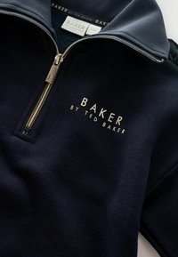 Námornícky zipsový sveter s vysokým golierom, zlatým zipsom a značkovým logom "BAKER by TED BAKER" v zlatom písme na hrudi. Mäkká textúra látky.