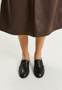 Mocassini in pelle nera con una texture liscia, design eleganti e punta affusolata. Posizionati su una gonna marrone scuro.