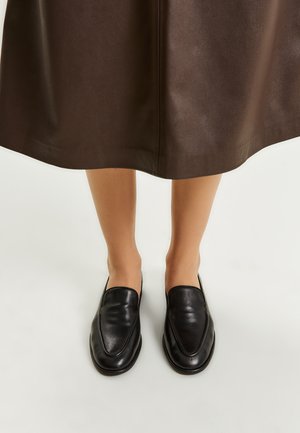 Black leather loafers with a smooth texture, sleek design, and pointed toe shape. Set against a dark brown skirt.
