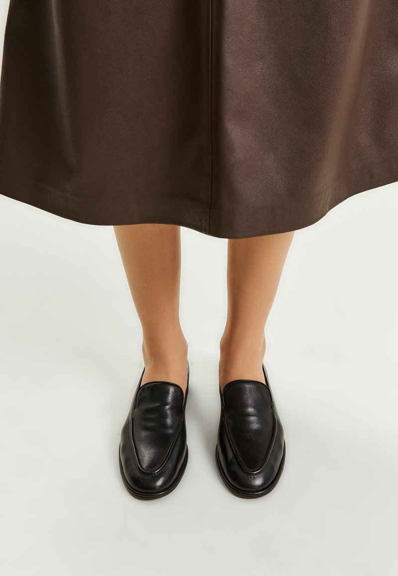Mocassini in pelle nera con una texture liscia, design eleganti e punta affusolata. Posizionati su una gonna marrone scuro.
