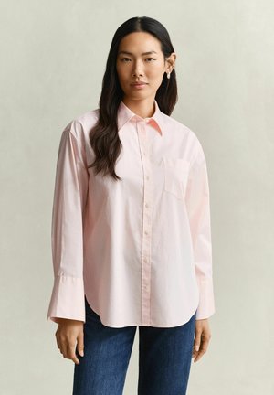 Jeune femme aux longs cheveux foncés portant une chemise rose clair boutonnée et un jean bleu sur un fond clair uni