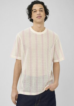 Jeune homme aux cheveux bouclés portant un t-shirt à manches courtes en maille rose clair et un jean foncé, debout devant un fond gris uni.