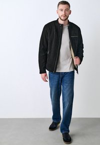 Sort ruskindsjakke med høj krave og lynlåslukning, båret over en lysegrå skjorte, matchet med blå jeans og sorte sko.