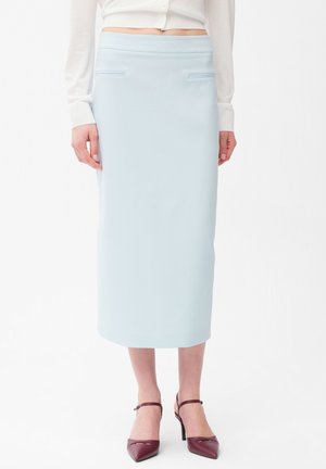 Kvinde iført en lyseblå midi pencil nederdel med skrålommer, kombineret med en hvid langærmet top og bordeaux spidse hæle.