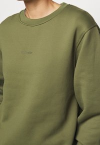 Olivgrön sweatshirt med rund halsringning och långa ärmar. Har en präglad logotyp på bröstet och ett slätt, texturerat tyg.