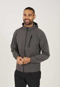 Grijze, lichte jas met een capuchon, een rits aan de voorkant, elastische manchetten en een ritszak op de borst. Soepele textuur en minimalistisch ontwerp.