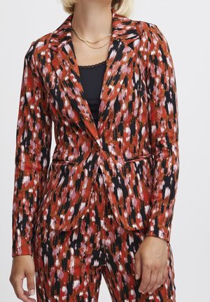 Kobieta ubrana w marynarkę i spodnie w abstrakcyjny wzór w kolorach czerwonym, czarnym i białym, z czarnym topem i złotymi łańcuszkami na szyję.