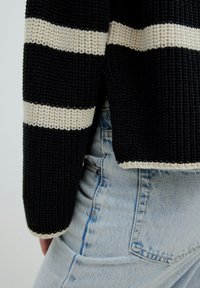 Pull en maille rayé noir et crème avec une texture côtelée, caractérisé par une coupe décontractée et un ourlet qui tombe juste au-dessus d'un jean en denim bleu clair.
