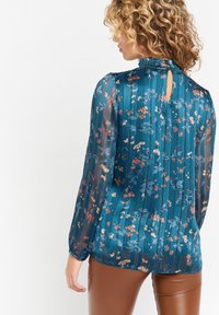 Tealblomstrad blus med volangkrage, genomskinliga långa ärmar och nyckelhålsrygg. Har ett strukturerat, vertikalt randigt mönster och blomsterdekorationer.