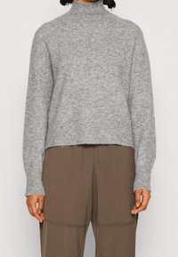 Grau gestrickter Rollkragenpullover mit gerippten Bündchen und Saum, der eine weiche Textur und ein kurzes Design aufweist. Getragen über braunen Hosen.