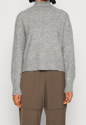 Pull en maille gris avec col roulé, avec des poignets et un bas côtelés, offrant une texture douce et un design court. Porté sur un pantalon marron.