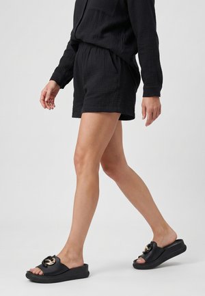 Person, die schwarze, strukturierte Shorts, ein passendes langärmeliges Oberteil und schwarze Slip-On-Sandalen mit goldener, runder Schnalle trägt.