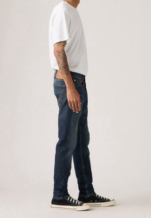 Mand iført en hvid t-shirt, mørkeblå jeans, sorte sneakers og synlige tatoveringer på venstre arm står sideværts.