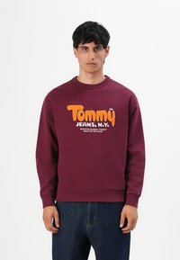 Hombre vistiendo un sudadera burdeos con el texto en naranja y blanco "Tommy Jeans, N.Y." y jeans azul oscuro, de pie contra un fondo blanco.