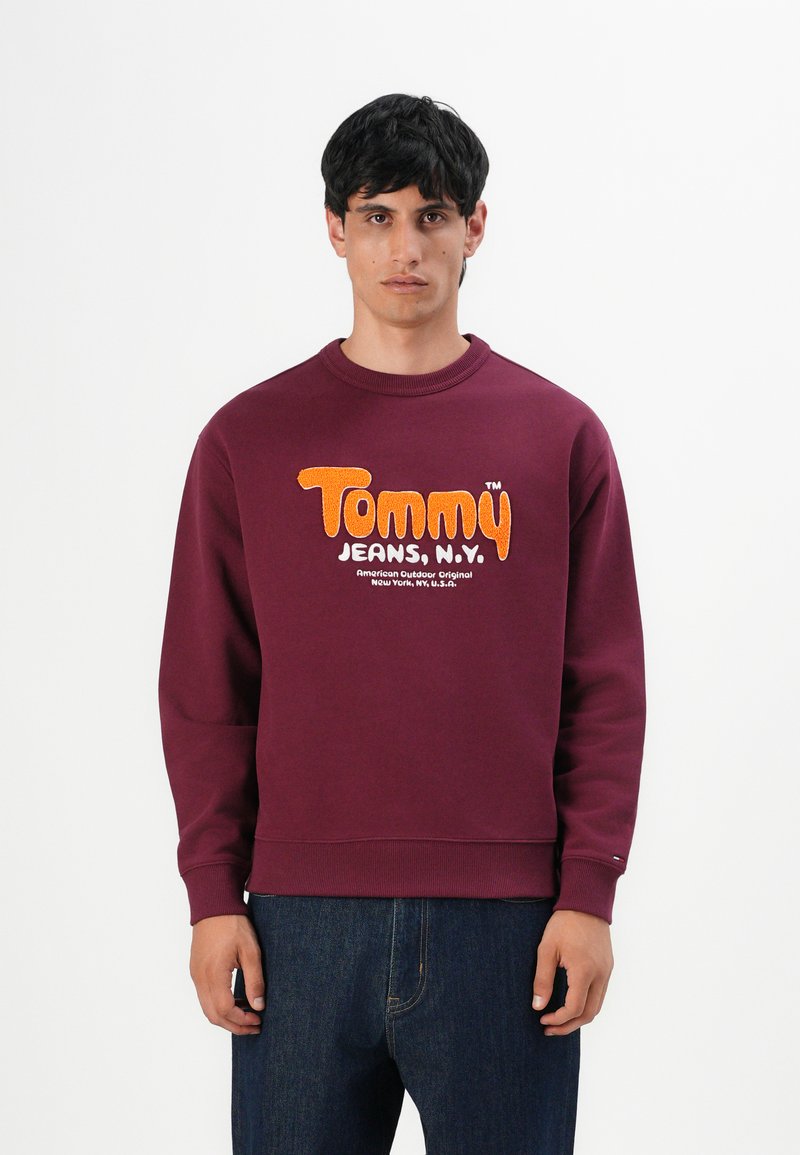 Hombre vistiendo un sudadera burdeos con el texto en naranja y blanco "Tommy Jeans, N.Y." y jeans azul oscuro, de pie contra un fondo blanco.