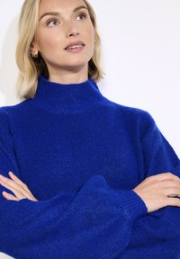 Blonde Frau mit heller Haut, die einen leuchtend blauen Pullover mit hohem Kragen trägt, Arme verschränkt, leicht nach oben und zur Seite blickend.