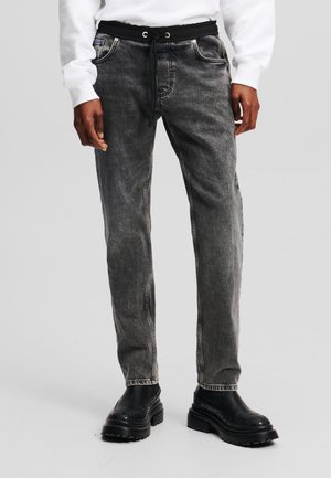 Volwassene draagt vervaagde zwarte spijkerbroek met een zwarte elastische tailleband en trekkoord, gecombineerd met zwarte stevige laarzen en een wit shirt met lange mouwen.