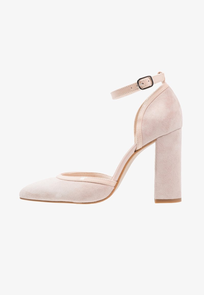 Escarpin en suède rose avec un bout pointu, une bride de cheville et un talon épais. Présente un liseré brillant le long de la bordure supérieure.