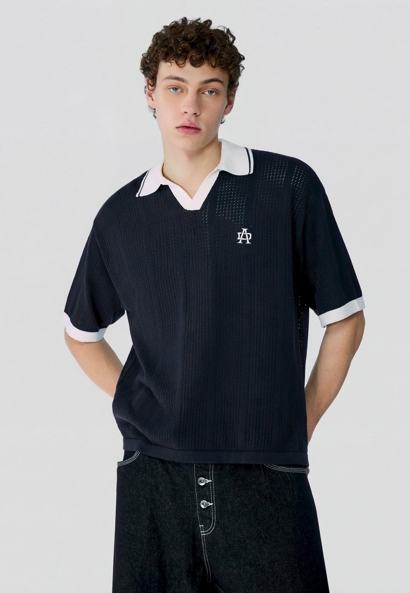 Giovane uomo con capelli ricci che indossa una polo in maglia blu navy con colletto e polsini bianchi, e pantaloni scuri con bottoni visibili.