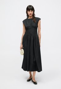 Czarna sukienka bez rękawów z marszczonym górnym partem, lejącym się dołem i wysokim dekoltem. Osoba nosząca ją trzyma małą, błyszczącą, beżową torebkę.