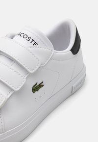 Fehér sneaker tépőzárral, zöld Lacoste logóval és fekete akcentussal a sarkán. Simító szintetikus anyag.