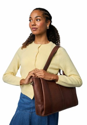 Hnědá kožená kabelka přes rameno s pevnou strukturou, zdobená vertikálními ševy a dlouhým popruhem. Modelka má na sobě žlutý svetr.