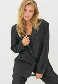 Black blazer featuring a textured check pattern, single button closure, and two front pockets. Long sleeves and a relaxed fit.