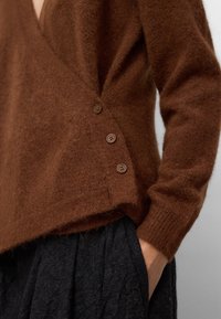 Brązowy, puszysty sweter z dekoltem w kształcie litery V, z trzema guzikami po boku i ściągaczami na rękawach. W zestawieniu z teksturowanym czarnym dołem.