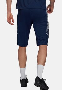 Hombre vestido con pantalones cortos deportivos azul marino con rayas laterales blancas, calcetines blancos y zapatillas deportivas negras, de pie con la espalda hacia el espectador.