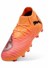Scarpa da calcio di un brillante arancione con superficie testurizzata, logo nero e dettagli rosa. Presenta un colletto aderente e una suola con tacchetti per garantire trazione.