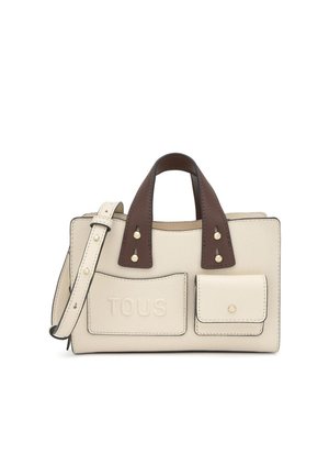Bolso de cuero crema con asas marrón oscuro, dos bolsillos delanteros, uno con relieve que dice "TOUS", y una correa ajustable sobre fondo blanco.