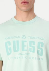 Camisa de algodón verde menta con cuello redondo y un estampado gráfico que incluye la palabra "GUESS" y elementos de texto en un tono contrastante.