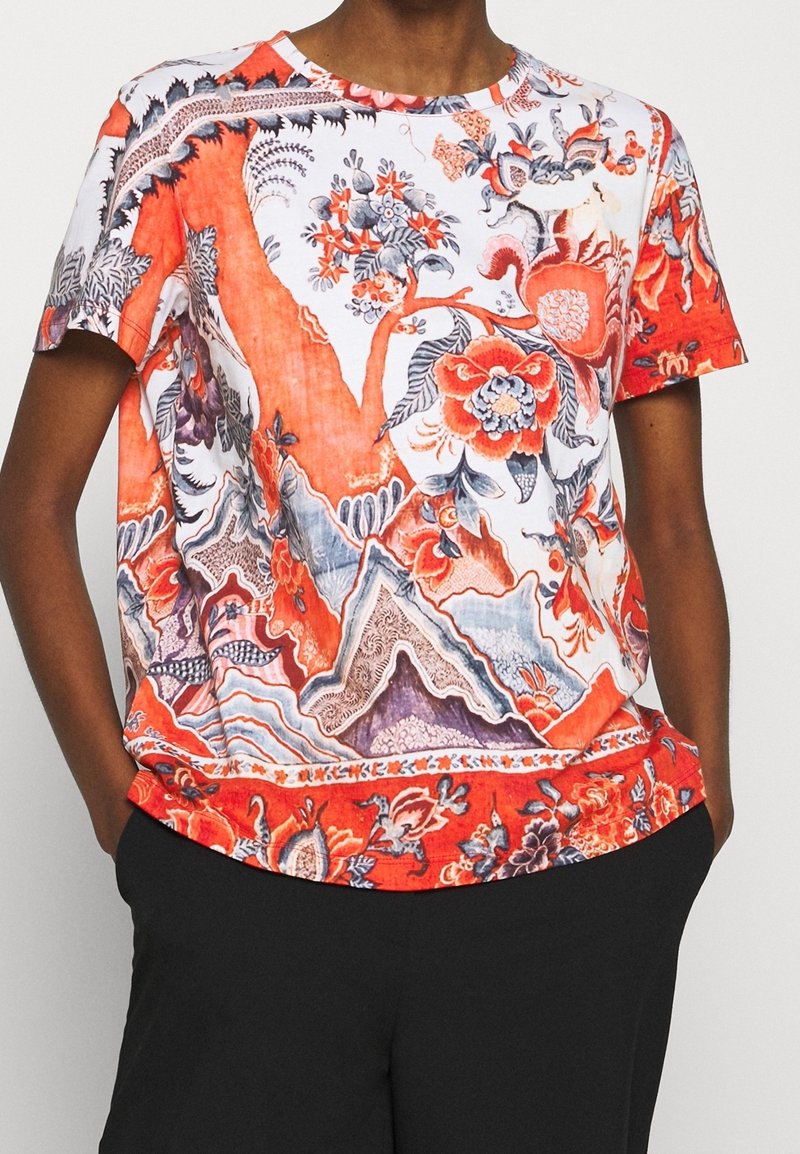 Chemise à manches courtes avec des motifs floraux et abstraits rouges, blancs et gris éclatants, portée avec un pantalon noir.